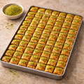 Baklava squares on a baking tray with a bowl of green pistachio on a beige surface